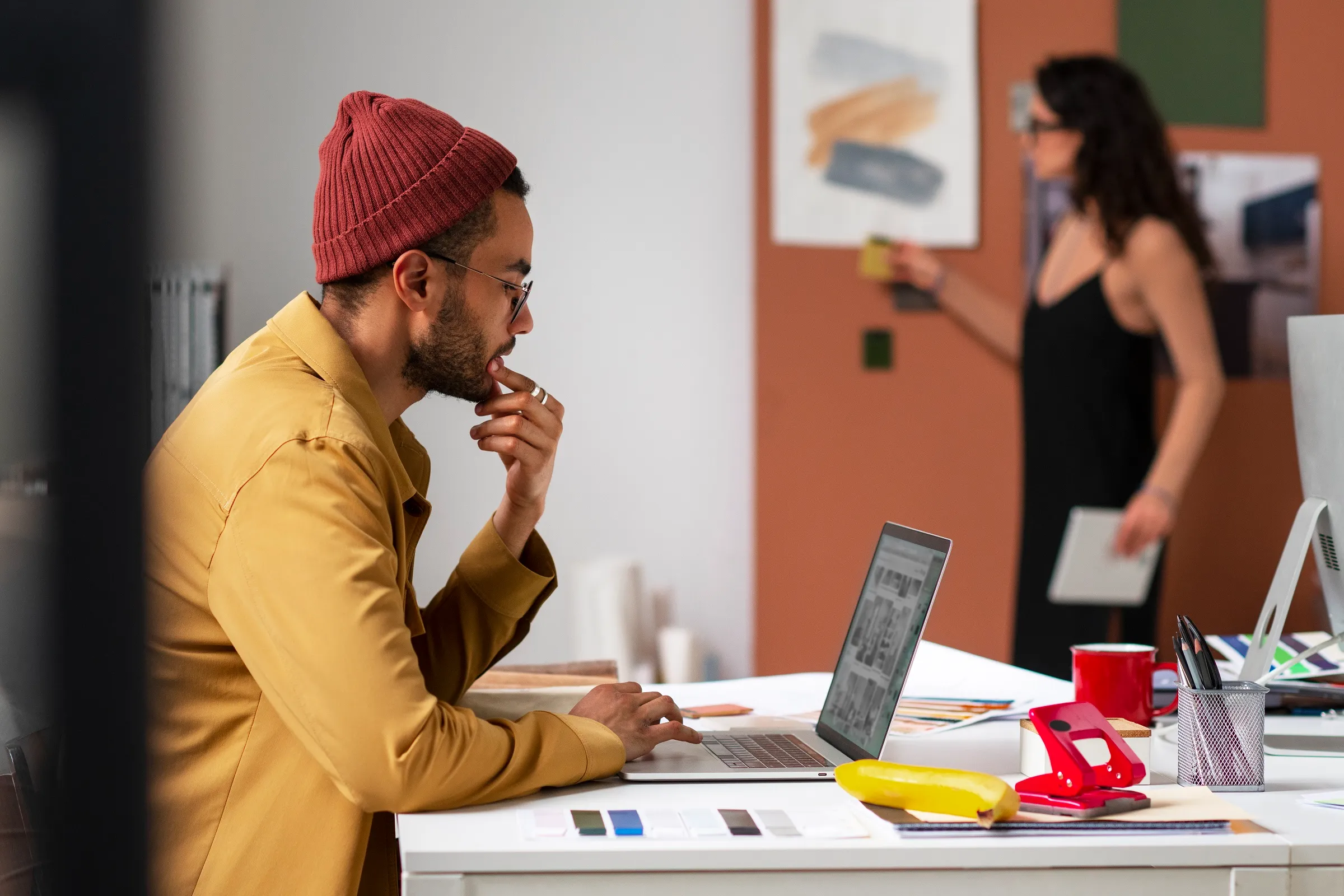 Male creative with red beanie working on his laptop, female colleague in the background